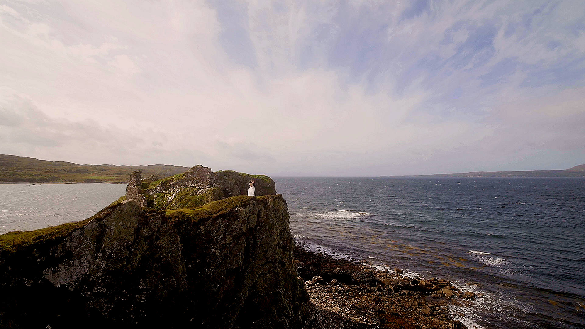 Wide-angle drone cinematography of a wedding couple standing on the sheer sea cliffs near the ruins of Dunscaith Castle, Isle of Skye.